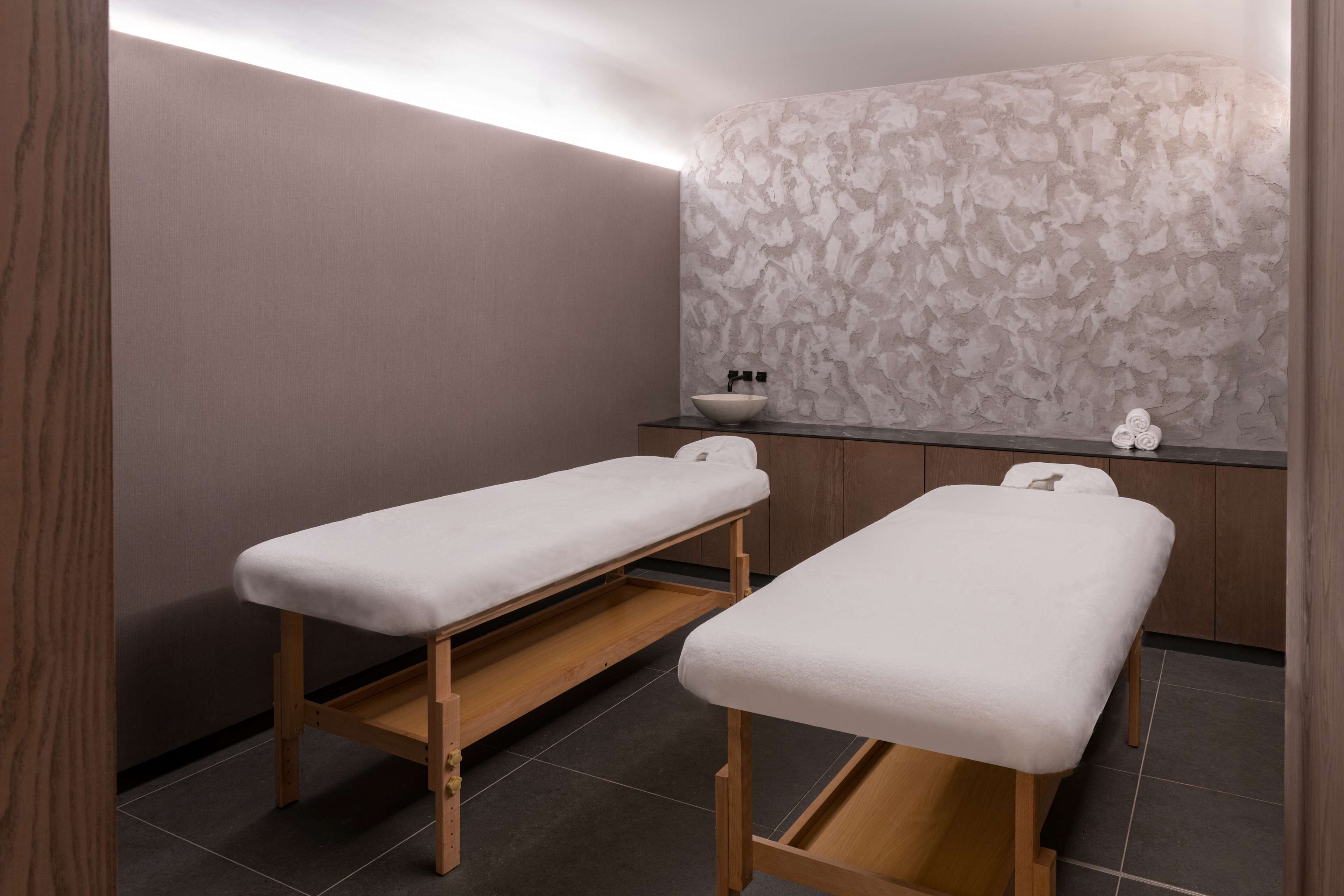 Ein moderner Spa-Raum mit zwei Holzbettliegen, einer strukturierten Wand und einer kleinen Theke mit Schale und Flaschen.