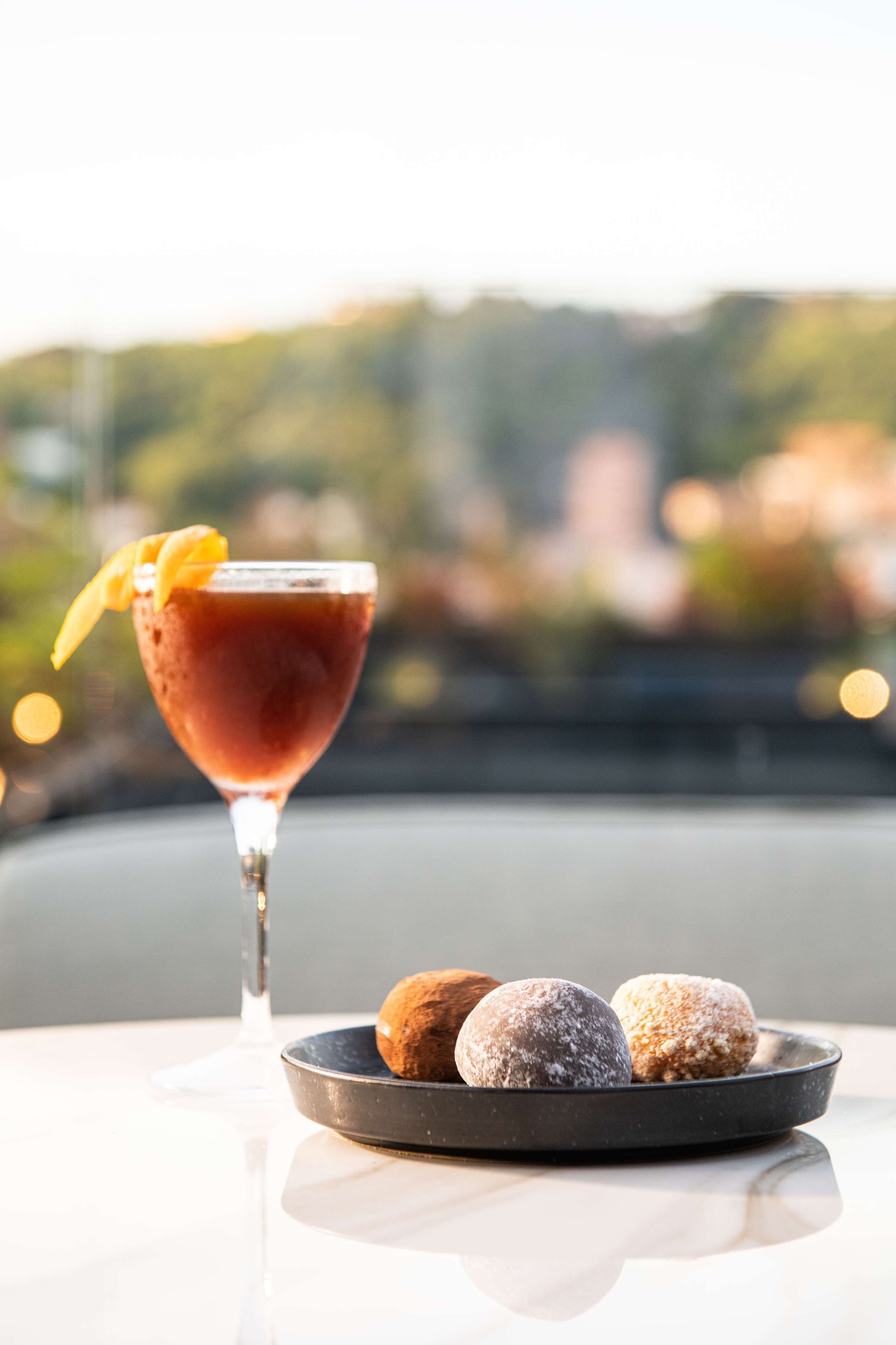 A martini glass with a garnish and a plate of three doughnuts on a table with a blurred outdoor view in the background.