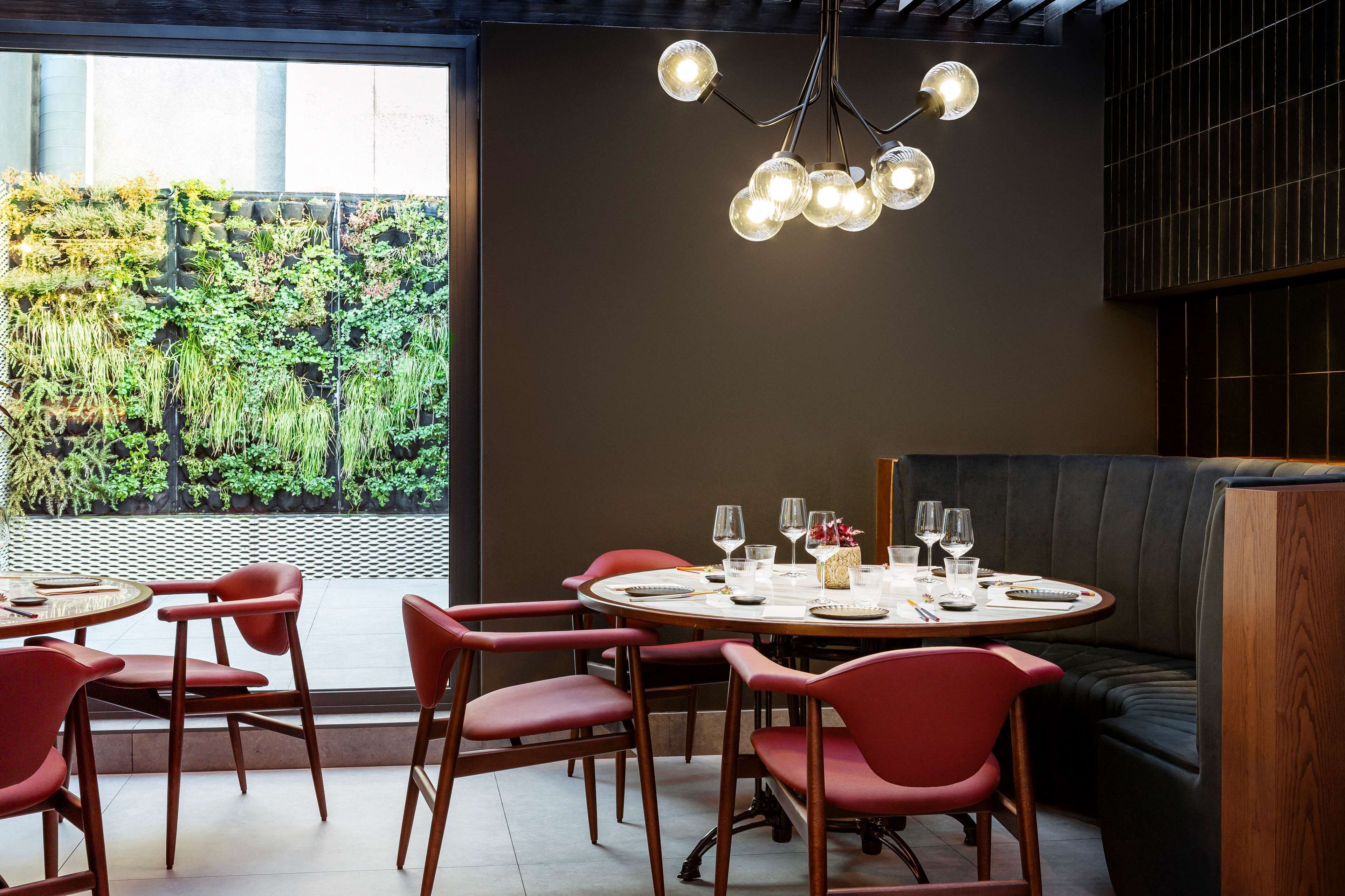 Een restaurant met ronde tafels, rode stoelen en een groene wand met planten.