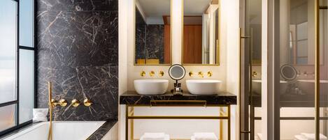 A modern bathroom with a black marble wall, a white bathtub, two sinks with gold fixtures, and a glass-enclosed shower area.