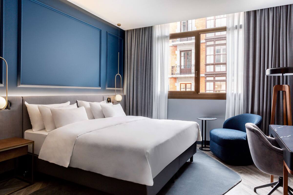 A hotel room with a large bed, a desk, a chair, and a view of a building through the window.