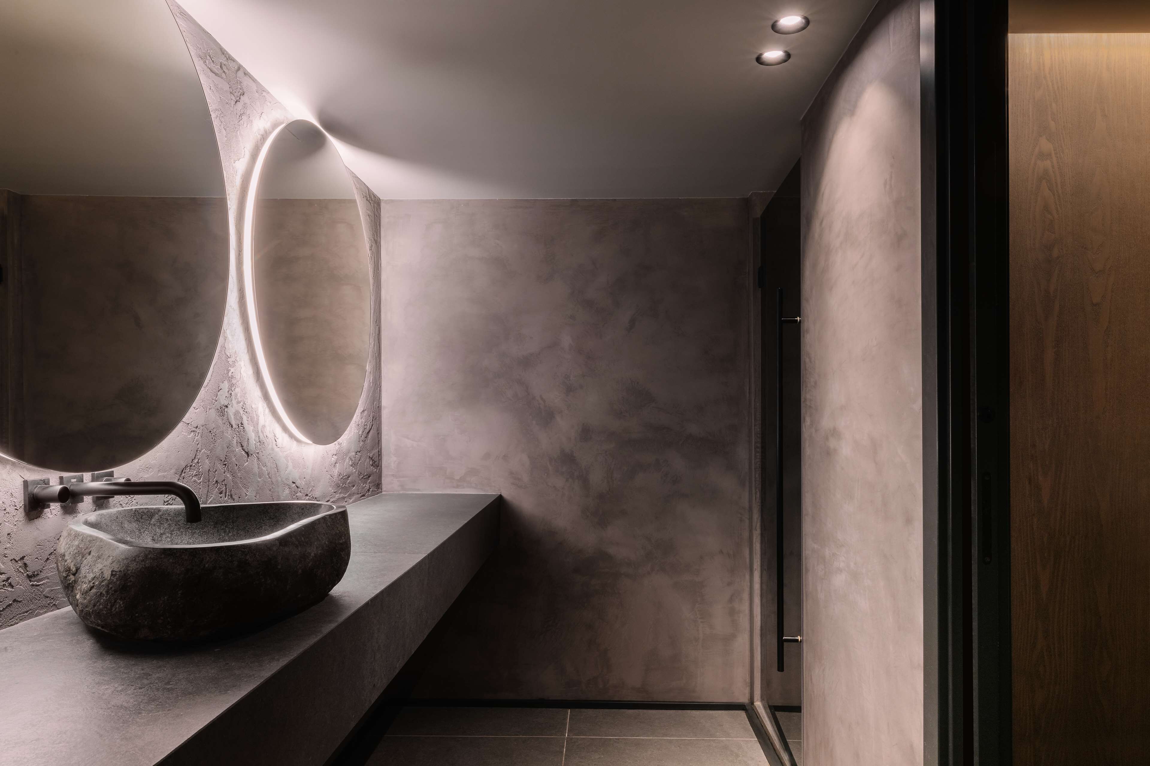 Un baño moderno con un espejo grande de forma ovalada, un lavamanos de piedra y un diseño minimalista.