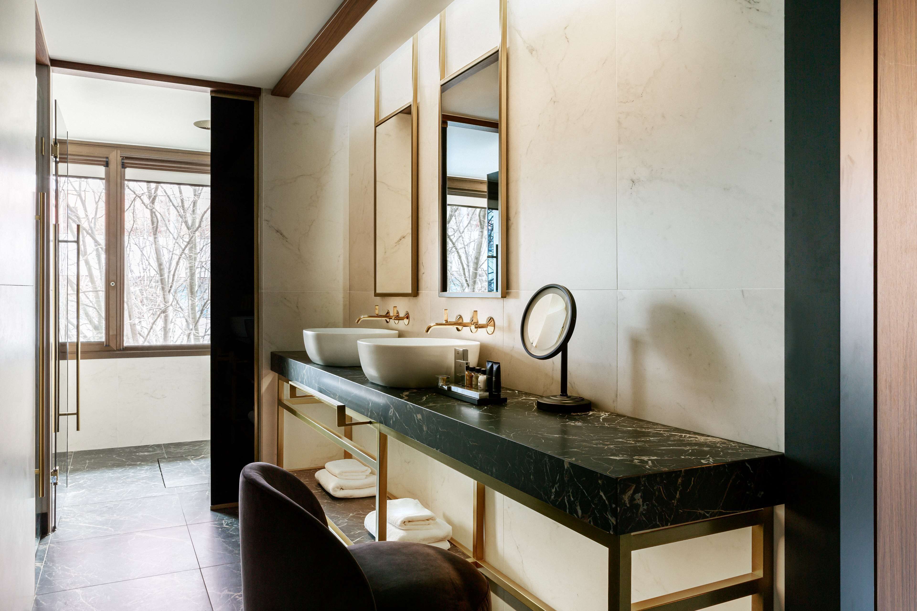 Une salle de bain moderne avec un comptoir en marbre, deux lavabos, un miroir et une vue sur les arbres par une fenêtre.