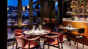A restaurant with round tables and red chairs, a bar area with a wooden counter, and a view of a cityscape at dusk. - Radisson Collection Bilbao (Bilbao)
