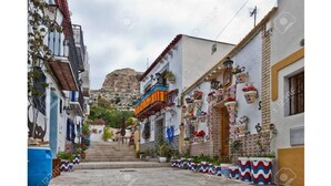 Una calle estrecha flanqueada por edificios, una escalera que sube hacia una colina y macetas con plantas al pie de la escalera.