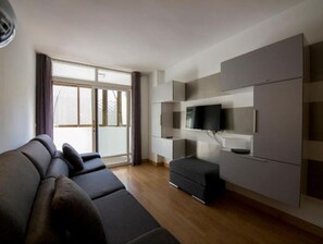 Un salon moderne avec un canapé d’angle, un téléviseur à écran plat fixé au mur et une porte-fenêtre coulissante donnant sur un balcon.