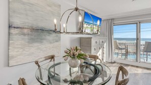 A modern dining area with a glass table, wooden chairs, a chandelier, and a view of the ocean through large windows.