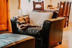 Un sillón de cuero con un edredón estampado, una mesita de madera, y en el fondo, una zona de comedor con sillas y una mesa de madera.