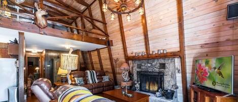 Interior de una cabaña de troncos con chimenea, sofá de cuero, vigas de madera y un trofeo de venado colgado.
