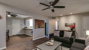 Ein modernes Wohnzimmer mit dunklem Holzboden, einem grauen Sofa mit gemusterten Kissen, einem Couchtisch und einem Deckenventilator.