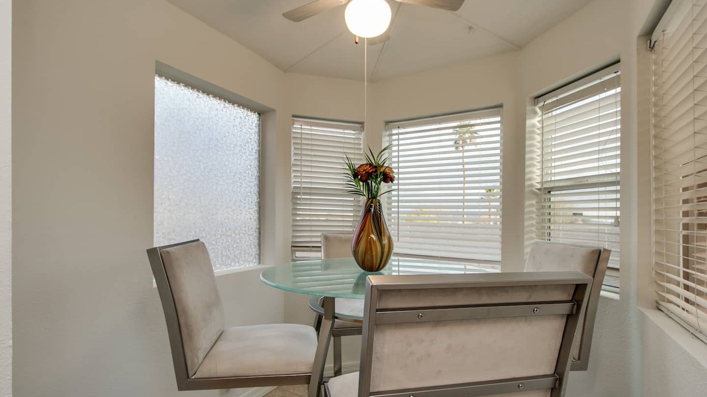 A dining area with a glass table, four chairs, a vase with flowers, and a ceiling fan.