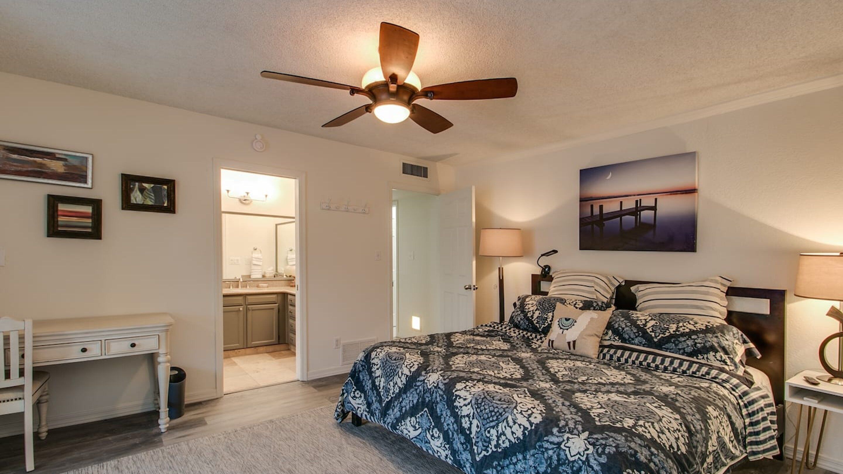 A bedroom with a bed, a desk, a ceiling fan, a bathroom, and a painting on the wall.