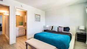 A bedroom with a bed, bedside tables, a bench, a bathroom with a sink and mirror, and a framed picture on the wall.