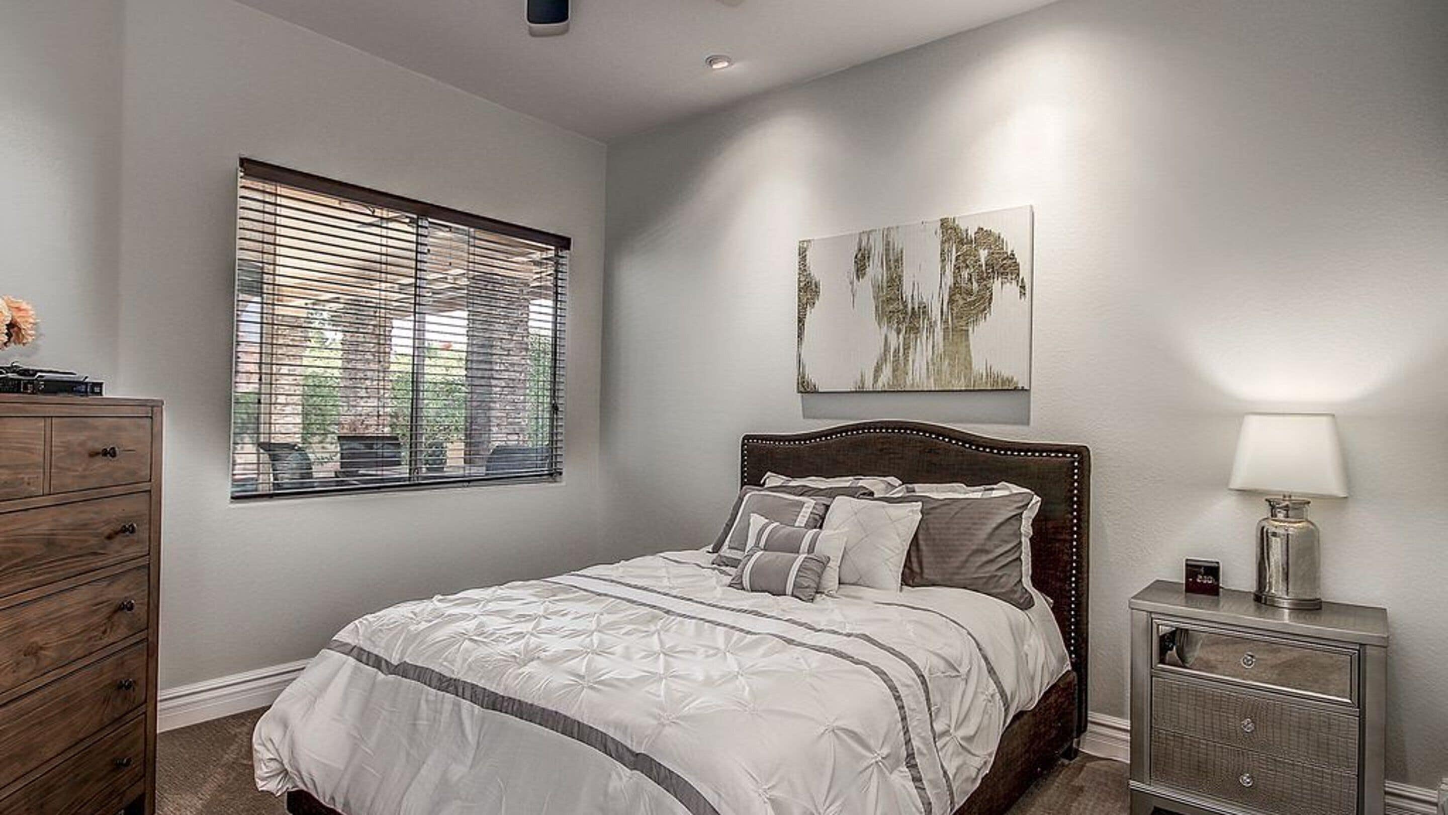 A bedroom with a bed, a nightstand, a dresser, a window with blinds, and a wall-mounted artwork.