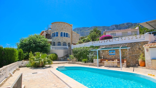 A swimming pool with stone tiles, a stone building with a tower, and a mountain in the background.