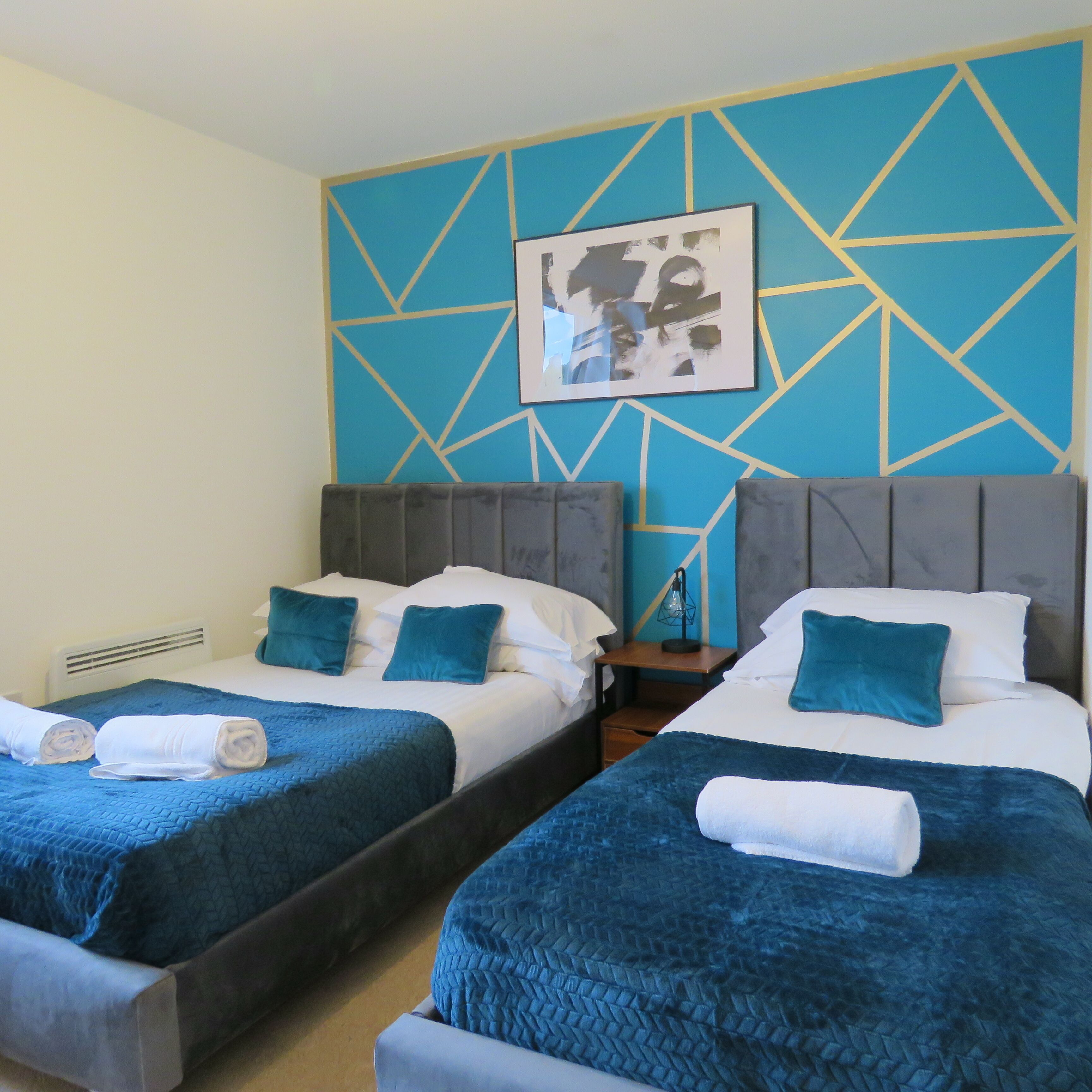 Une chambre d’hôtel avec deux lits, chacun recouvert d’une courtepointe matelassée bleue et agrémenté d’oreillers assortis. Un tableau abstrait encadré est accroché au mur, au-dessus des lits.