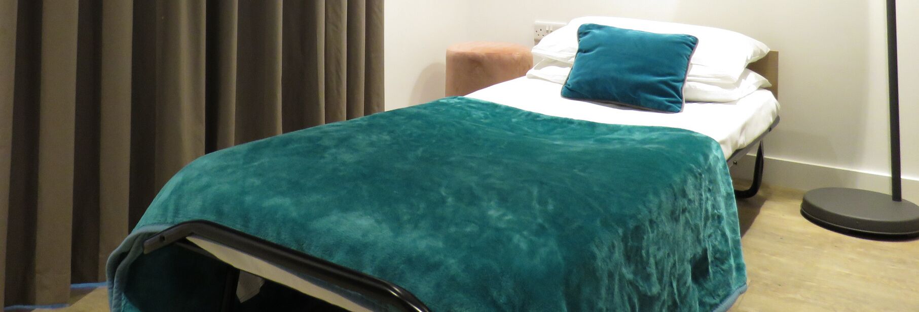 A neatly made bed with a teal blanket, a white pillow, and a blue pillow.