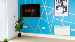 A room with a blue geometric wall design, a mounted flat-screen TV displaying "NETFLIX", a potted plant, a table with a magazine, and a white air conditioning unit. - Tudors eSuites Two Bedroom & Balcony (Birmingham)