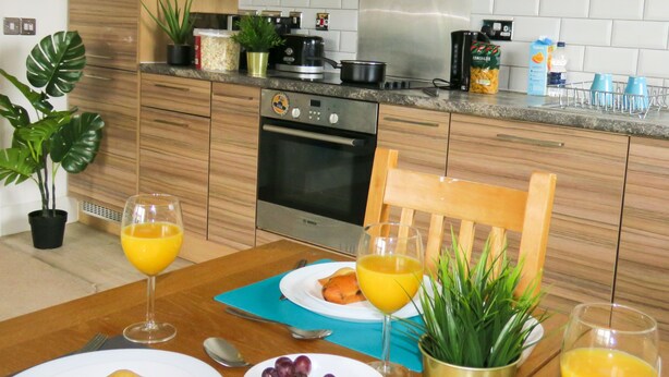 A modern kitchen with wooden cabinets, a Bosch oven, and a dining table set with plates, glasses, and a bowl of grapes. - Tudors eSuites Two Bedroom & Balcony (Birmingham)