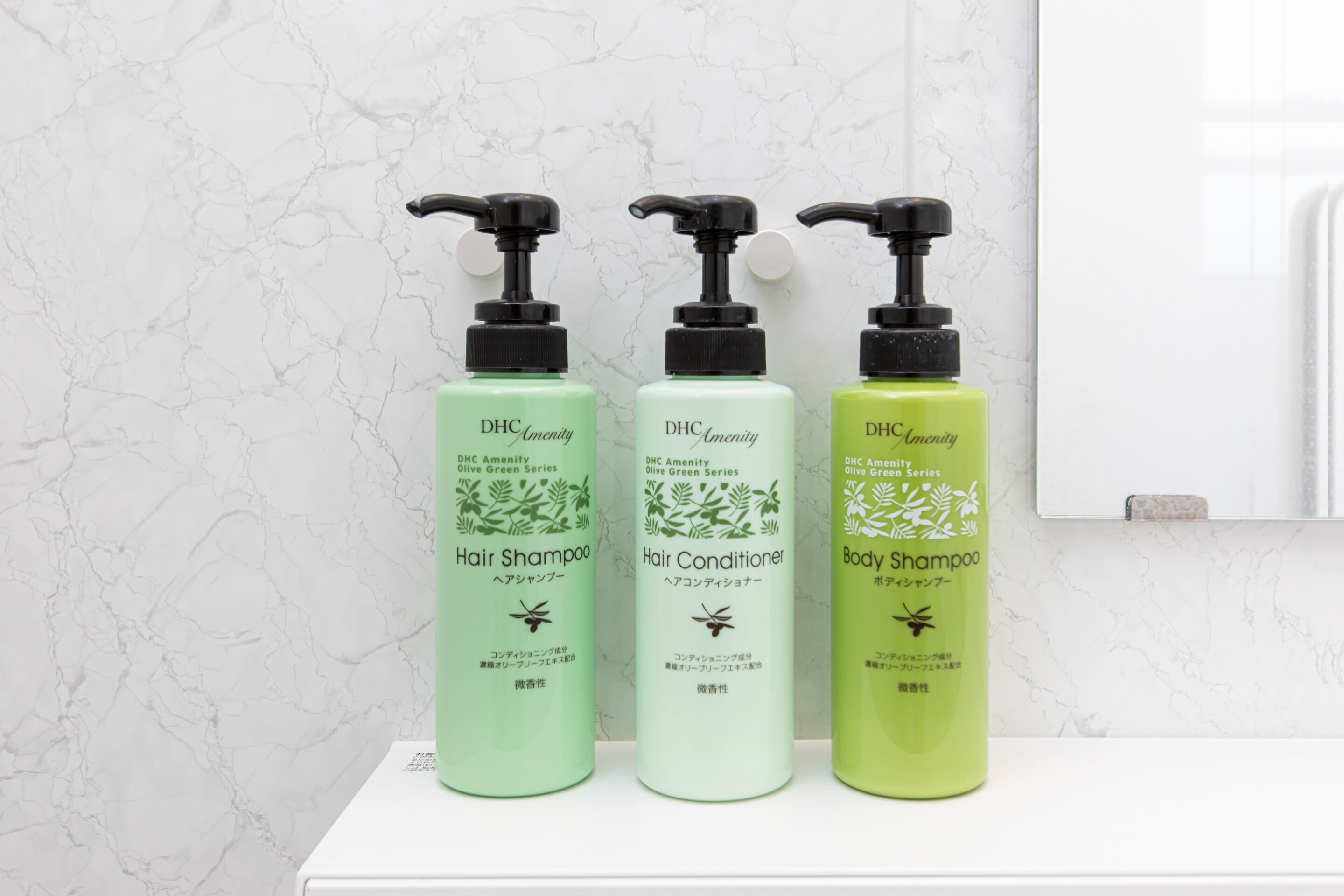 Three bottles of DHC Amenity Olive Green Series hair and body care products on a white shelf against a marble wall.