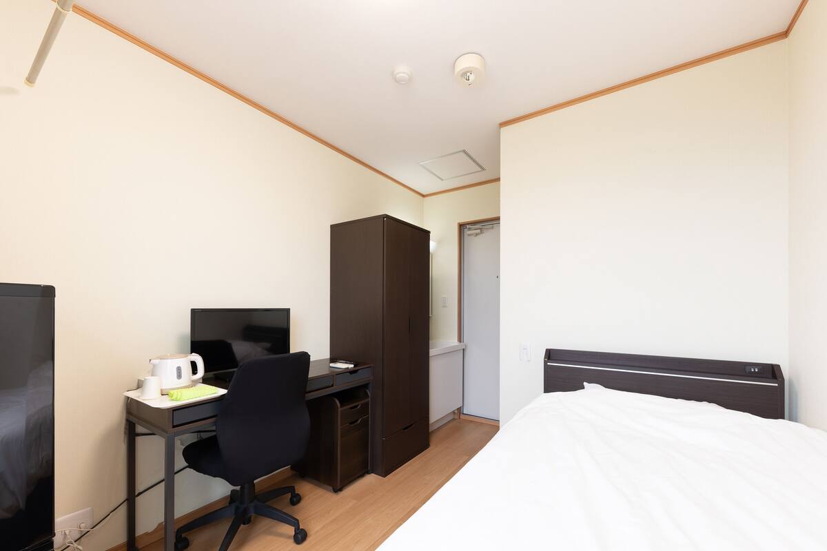 A hotel room with a bed, a desk with a computer, a chair, a wardrobe, and a television.