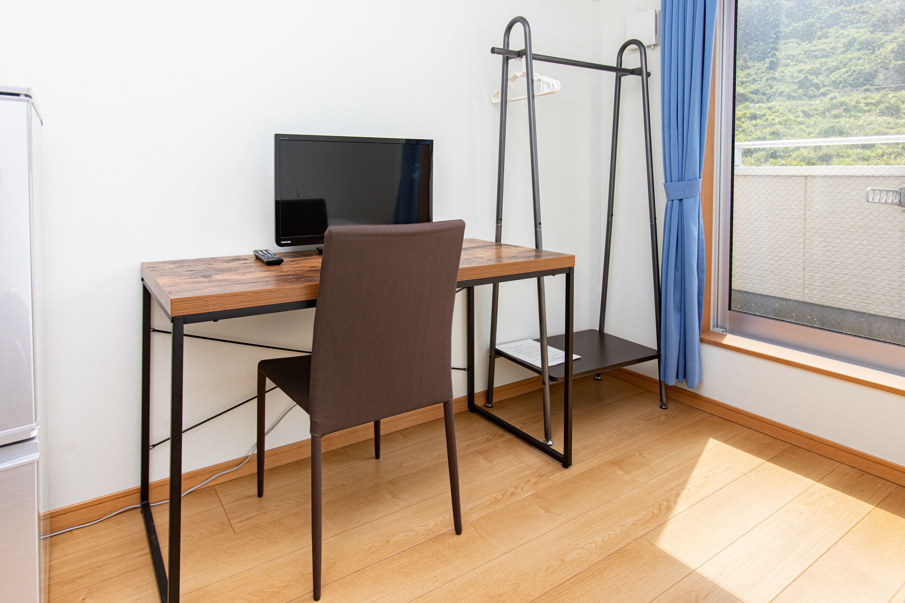 A room with a wooden desk, a chair, a television, and a window with a view of greenery.