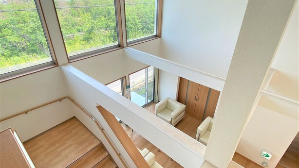 Un interior moderno con una escalera de madera, una zona de estar con sofá y sillón, y grandes ventanales que dan a zonas verdes y una masa de agua.
