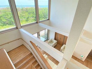 A modern interior with a wooden staircase, a seating area with a sofa and armchair, and large windows offering a view of greenery and a body of water. - Tabist Hotel Takagiya Iwaki (Iwaki)