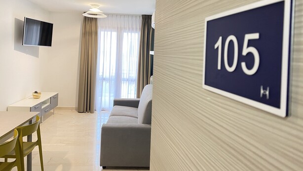 A modern hotel room with a sofa, a TV mounted on the wall, and a sign indicating room number 105. - APPARTAMENTO "FORGITELLA" 6 POSTI LETTO (Balestrate)