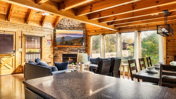 Interior acogedor de una cabaña con chimenea de piedra, vigas de madera, pared de piedra, televisor grande y zona de comedor con una isla de granito.