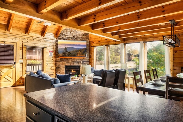 A cozy cabin interior with a stone fireplace, wooden beams, a stone wall, a large TV, and a dining area with a granite island.