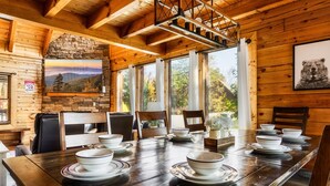 Interior de una cabaña de madera con una mesa de comedor para cuatro personas, una chimenea de piedra y un ventanal con vistas a la montaña.