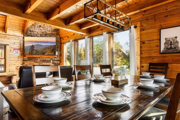 Interior de una cabaña de madera con una mesa de comedor para cuatro personas, una chimenea de piedra y un ventanal con vistas a la montaña.