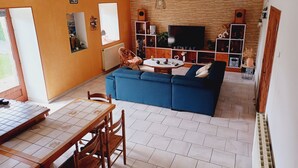 A living room with a blue sofa, a TV, and a dining area with a table and chairs. - Maison Dans une Ancienne Ferme sur 3,5ha (Plougonver)