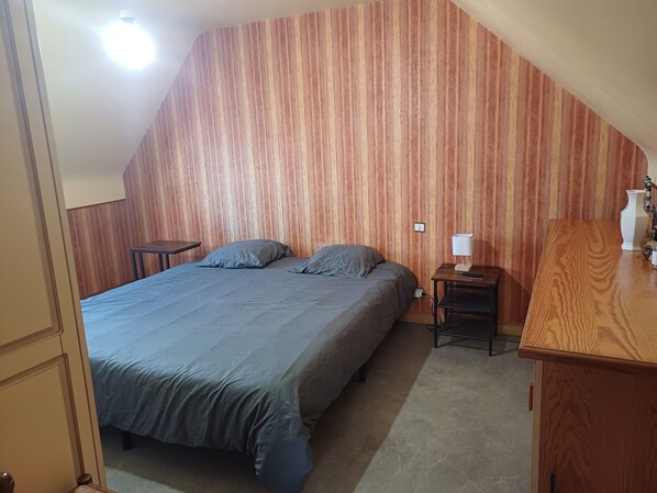 Un dormitorio con techo inclinado, una cama con ropa de cama gris, una mesita de noche con lámpara y un aparador de madera.