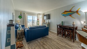 A living room with a blue sofa, wooden dining table, and a large fish mural on the wall. - Charming Ocean-view Condo at Ocean Pier 3 (North Myrtle Beach)