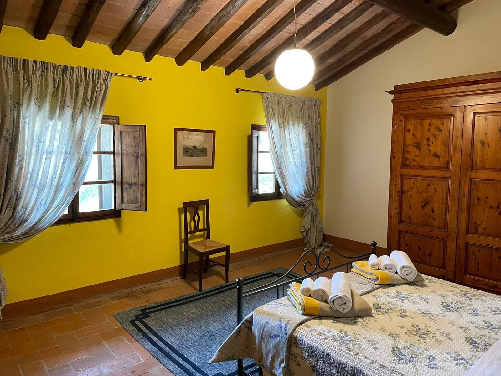 A room with yellow walls, wooden ceiling beams, a bed with rolled towels, a chair, and a window with curtains.