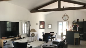 A spacious living room with exposed wooden beams, a large flat-screen TV, modern furniture, and a kitchen area in the background.