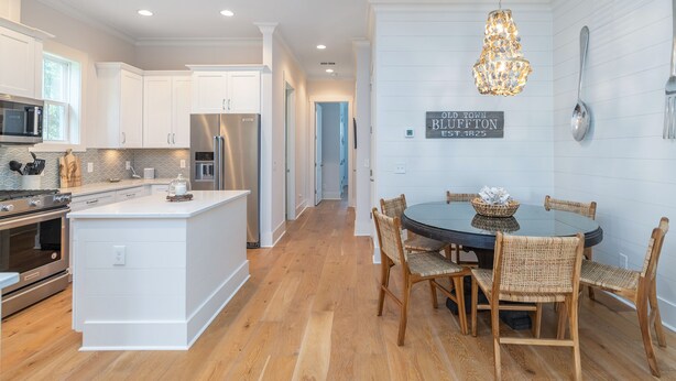 A modern kitchen with white cabinetry, a central island, and a dining area with wicker chairs. - Old Town Bluffton Home + FREE Golf Cart Luxury Home - Sleeps 8 (Bluffton)