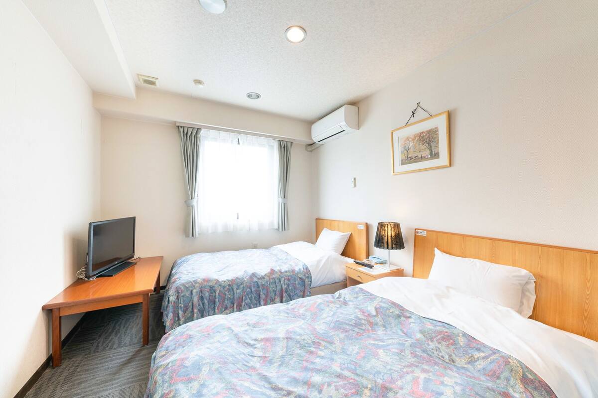 Una habitación de hotel con dos camas, un escritorio con televisor, una mesita de noche con lámpara y una ventana con cortinas.
