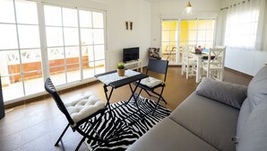 Ein geräumiges Wohnzimmer mit einer Couch, einem zebra-gemusterten Teppich, einem Esstisch mit Stühlen, einem Fernseher auf einem Schrank und einem Balkon mit großen Fenstern.