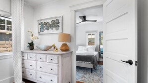 Een slaapkamer met een witte dressoir, een bed met witte lakens, een plafondventilator en een raam met uitzicht op bomen.