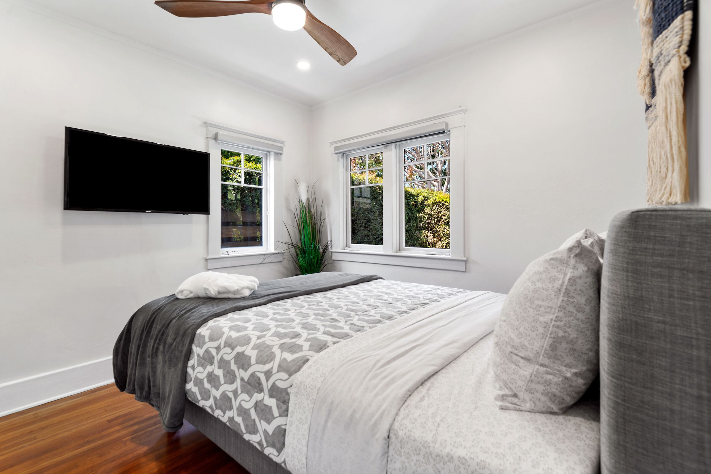 A bedroom with a bed, a ceiling fan, a flat-screen TV, and two windows with a view of greenery.