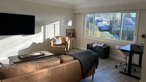A living room with a leather sofa, a flat-screen TV, a desk with a computer, and a large window offering a view of the neighborhood.