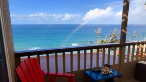 Un balcon avec une chaise rouge, une petite table et une vue sur l’océan, avec un arc-en-ciel en arrière-plan.