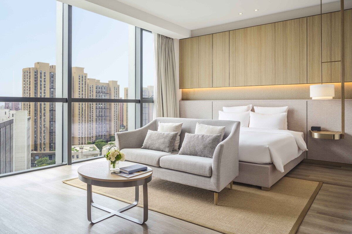 A modern hotel room with a large window, a sofa, a bed, and a small table with a vase of flowers.