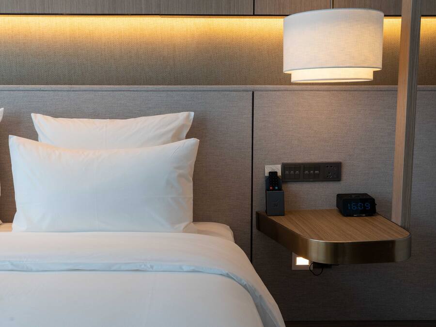 A neatly made bed with white linens, a bedside table with a digital clock, and a wall-mounted lamp.