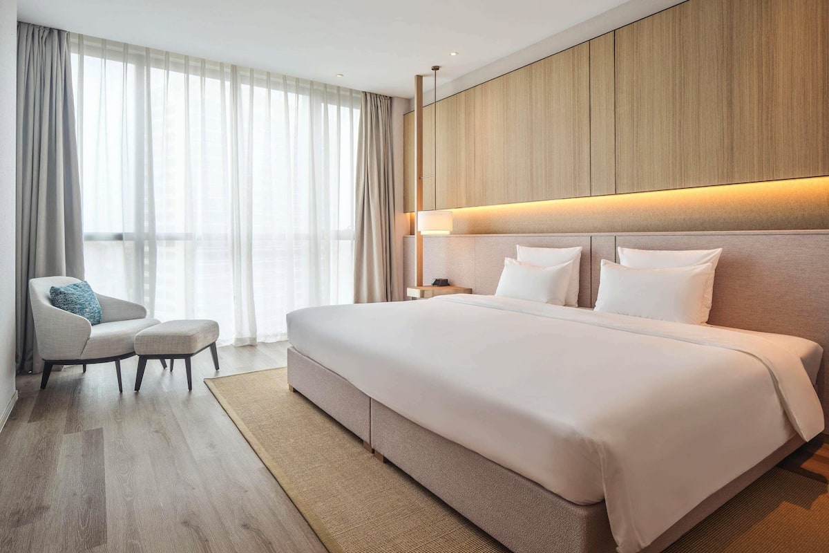 Ett modernt hotellrum med en stor säng, en stol och utsikt över staden genom tunna gardiner.