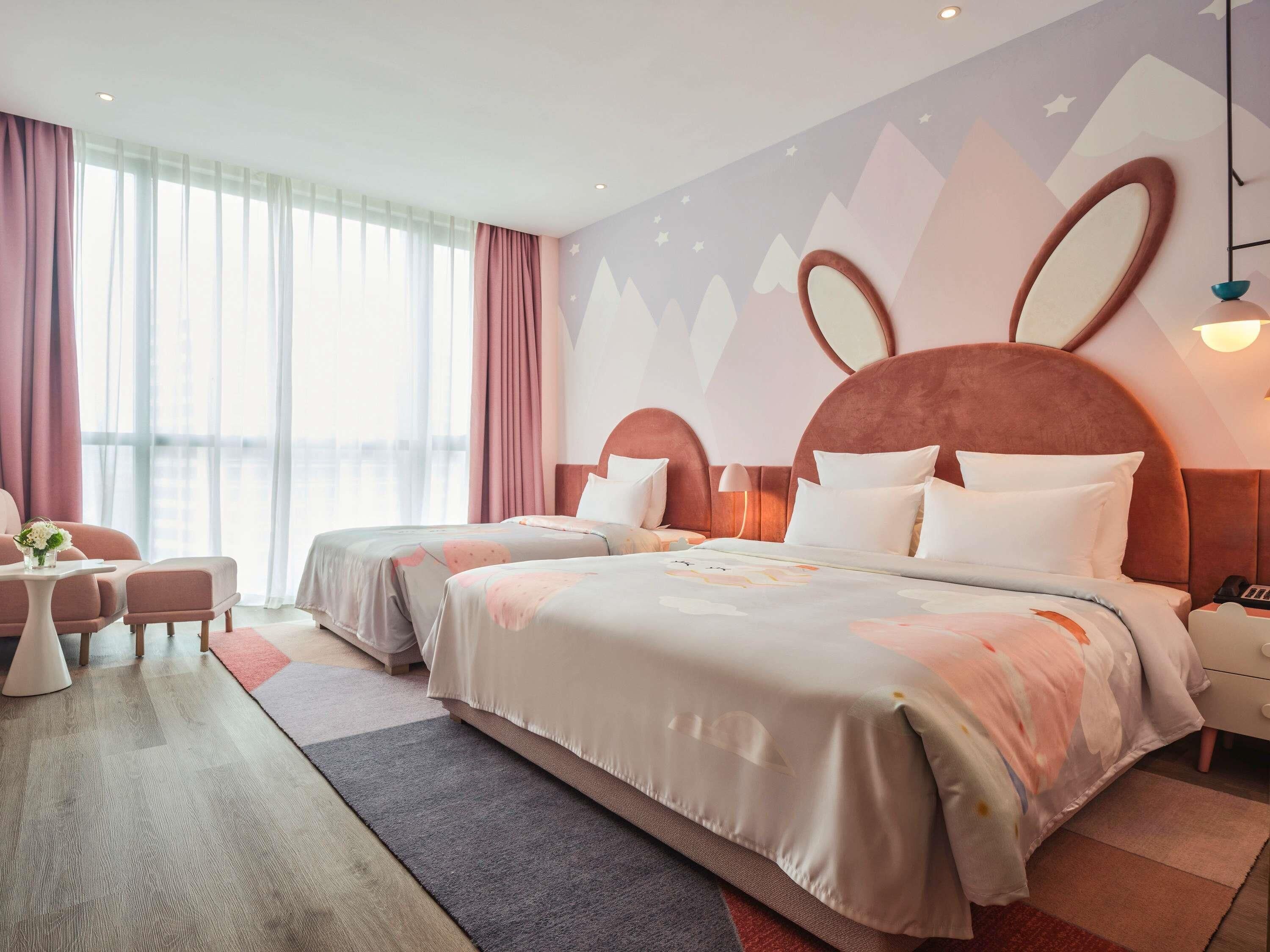 Ett hotellrum med två sängar, ett skrivbord, en stol och ett stort fönster med gardiner.