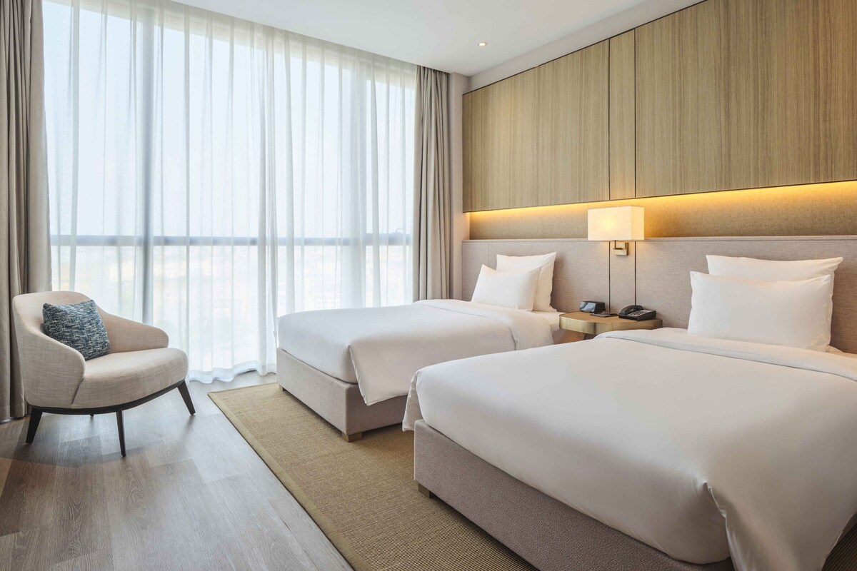 A hotel room with two beds, a chair, a nightstand, and a large window with sheer curtains.
